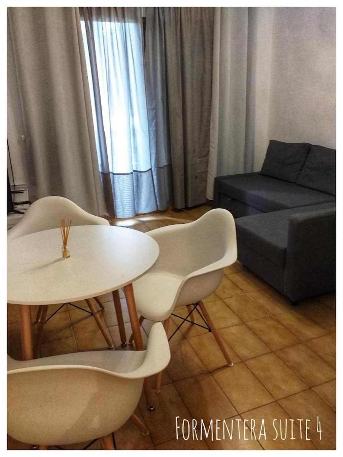 A light-filled living area features a round dining table surrounded by four white chairs. A comfortable gray sofa is positioned nearby, while sheer curtains gently diffuse natural light from a window.