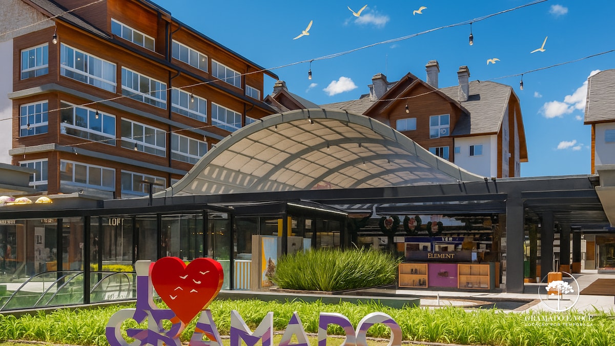 The image displays the entrance area of the Vita Boulevard complex, featuring a large colorful sign that reads 'I Love Gramado.' Modern buildings are visible in the background, complemented by a clear blue sky and decorative lighting strung overhead. Lush greenery adds a natural element to the setting.