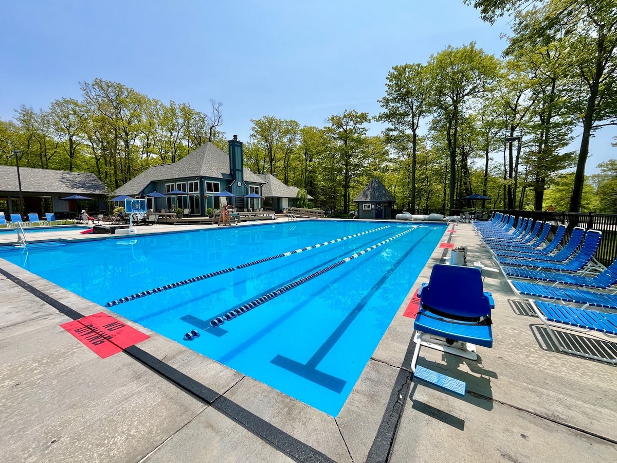 An expansive outdoor pool is set against a backdrop of trees, with bright blue water reflecting sunlight. Surrounding lounge chairs provide ample seating. A club house with a sloped roof is visible nearby, adding to the recreational atmosphere.