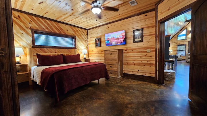 Bedroom with King bed, wall-mounted TV, pine plank walls, and ceiling fan — cozy and cool, single-story steps away from the action.