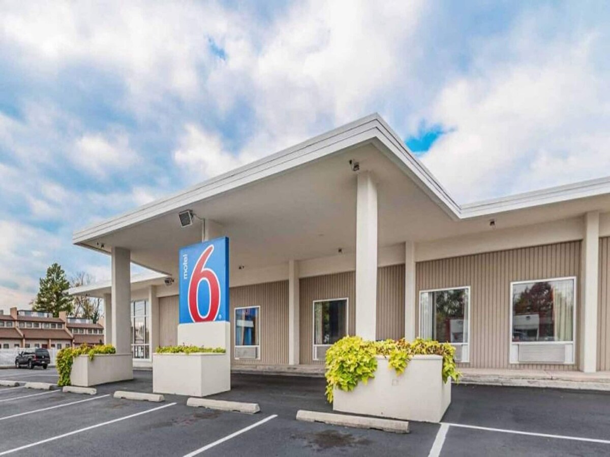 The exterior of a modern motel is depicted, featuring a covered entrance and a prominent blue sign displaying the number six. Large windows allow natural light to enter, while planters with greenery add a touch of color. A clean parking area is visible in front.