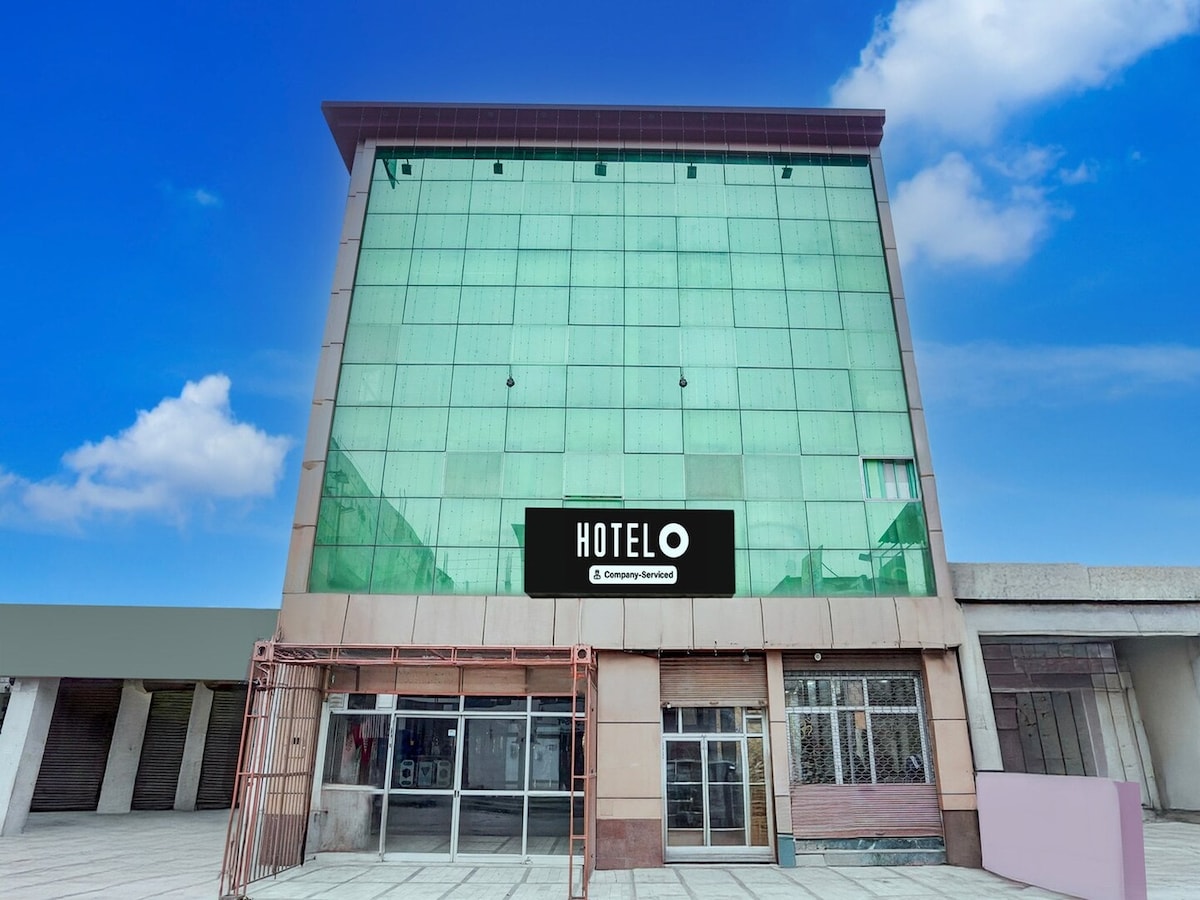 The hotel exterior is characterized by a modern glass façade with a large sign displaying the hotel name. The building features a spacious entrance area with a combination of glass and stone materials, complementing the clear blue sky backdrop.