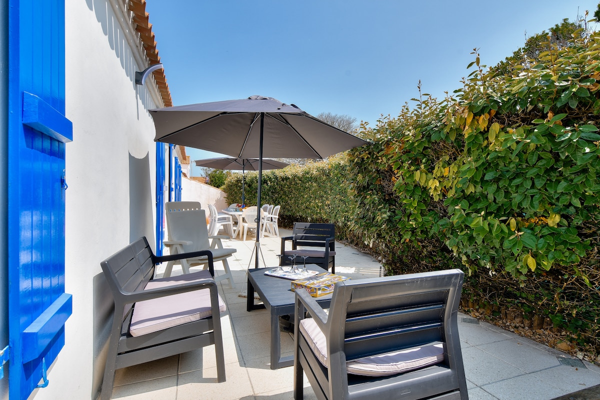 An outdoor terrace is featured, equipped with a table and seating, under a large umbrella for shade. Lush greenery lines the space, providing privacy. The area is designed for relaxation and outdoor dining, complementing the coastal ambiance of the home.