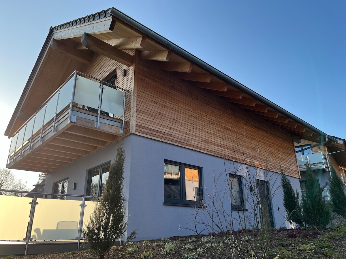 An exterior view of the chalet presents a modern design, featuring a sloped wooden roof and a spacious balcony with glass railings. The structure is complemented by landscaped greenery, with multiple windows reflecting natural light.