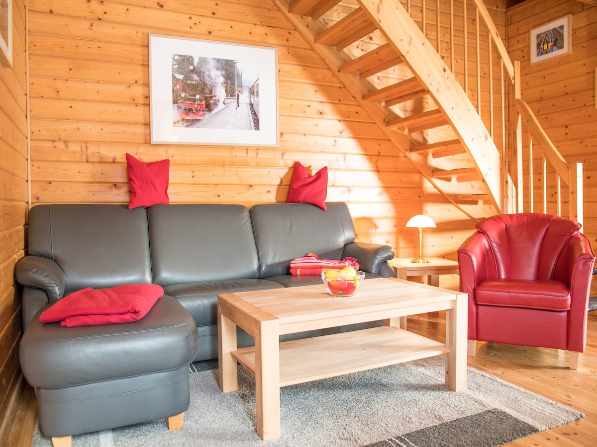 A wooden interior features a comfortable black leather sofa accompanied by a red armchair. A light wooden coffee table sits at the center. A staircase in the background leads to the upper level, while a framed image is displayed on the wall.