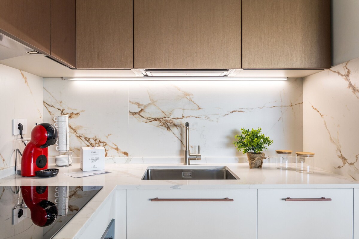 The kitchen features sleek cabinetry with a marble backsplash, complemented by modern appliances. A red coffee machine and a small plant add color to the bright space. The stainless steel sink is positioned below under-cabinet lighting, enhancing visibility and functionality.