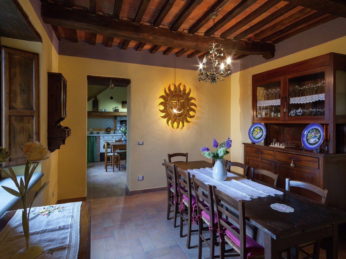 A spacious dining area features a large wooden table surrounded by eight chairs. Natural light filters through windows adorned with wooden frames. A decorative sun-shaped wall piece is mounted against a warm yellow backdrop, while a sideboard displays plates and glassware.