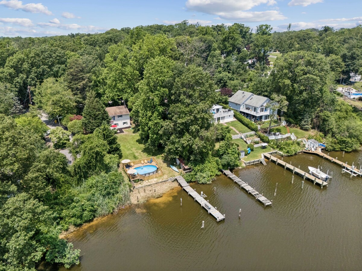 An aerial view captures the charming waterfront home surrounded by lush greenery. A private pool is visible in the spacious yard, along with a dock extending into the peaceful water, ideal for water activities. Nearby houses complement the serene landscape.