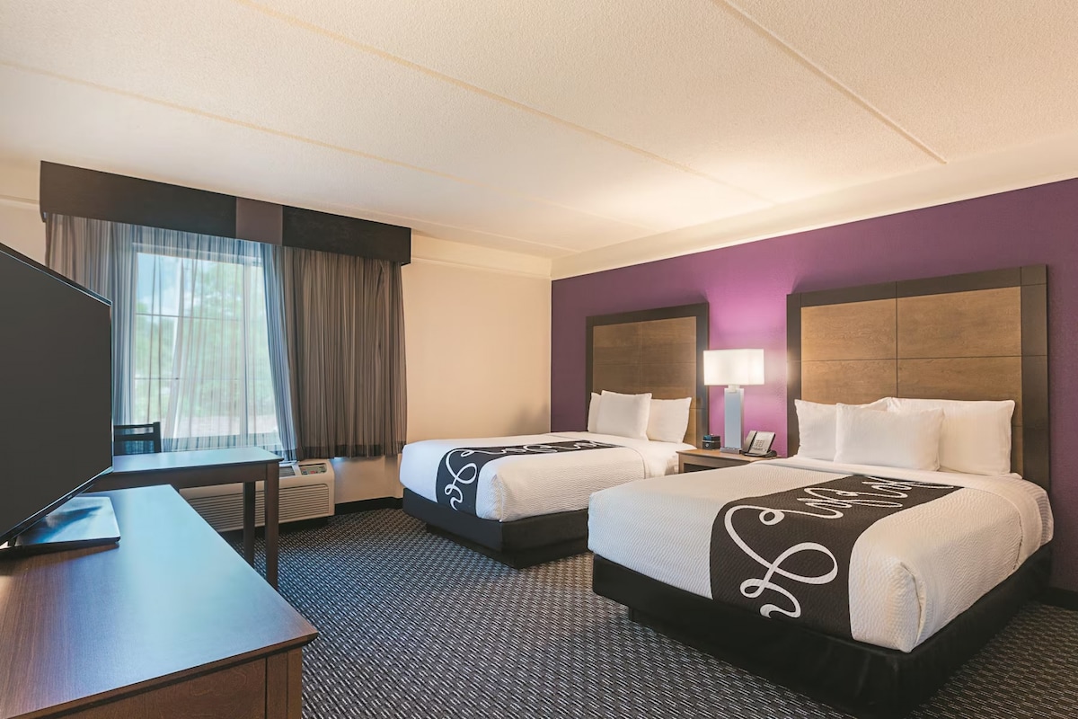 Dos camas con pared púrpura, ventana grande, escritorio y TV de pantalla plana