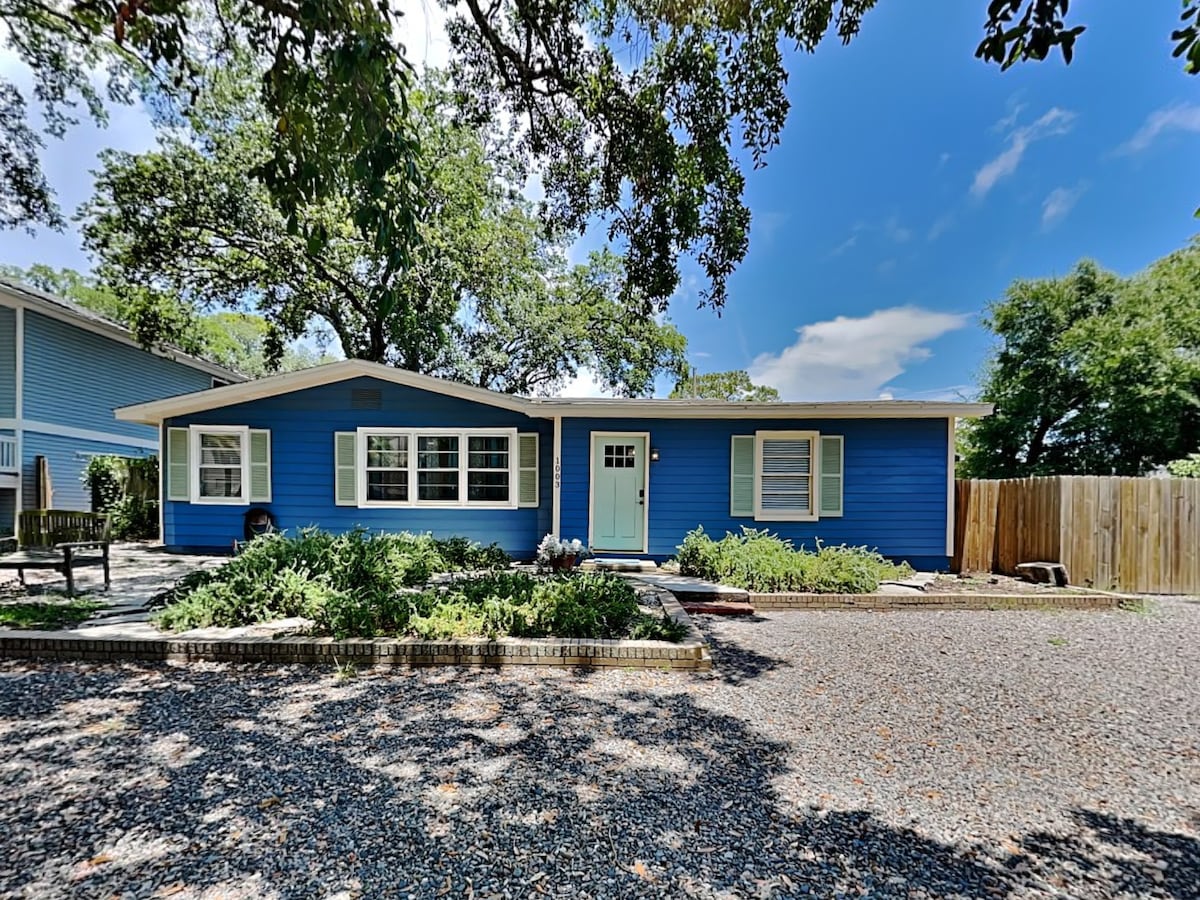 A charming blue single-story house is nestled among lush landscaping and gravel paths. The front porch features a light blue door, framed by ample windows that invite natural light. Nearby, comfortable outdoor seating is surrounded by greenery, creating a welcoming atmosphere.