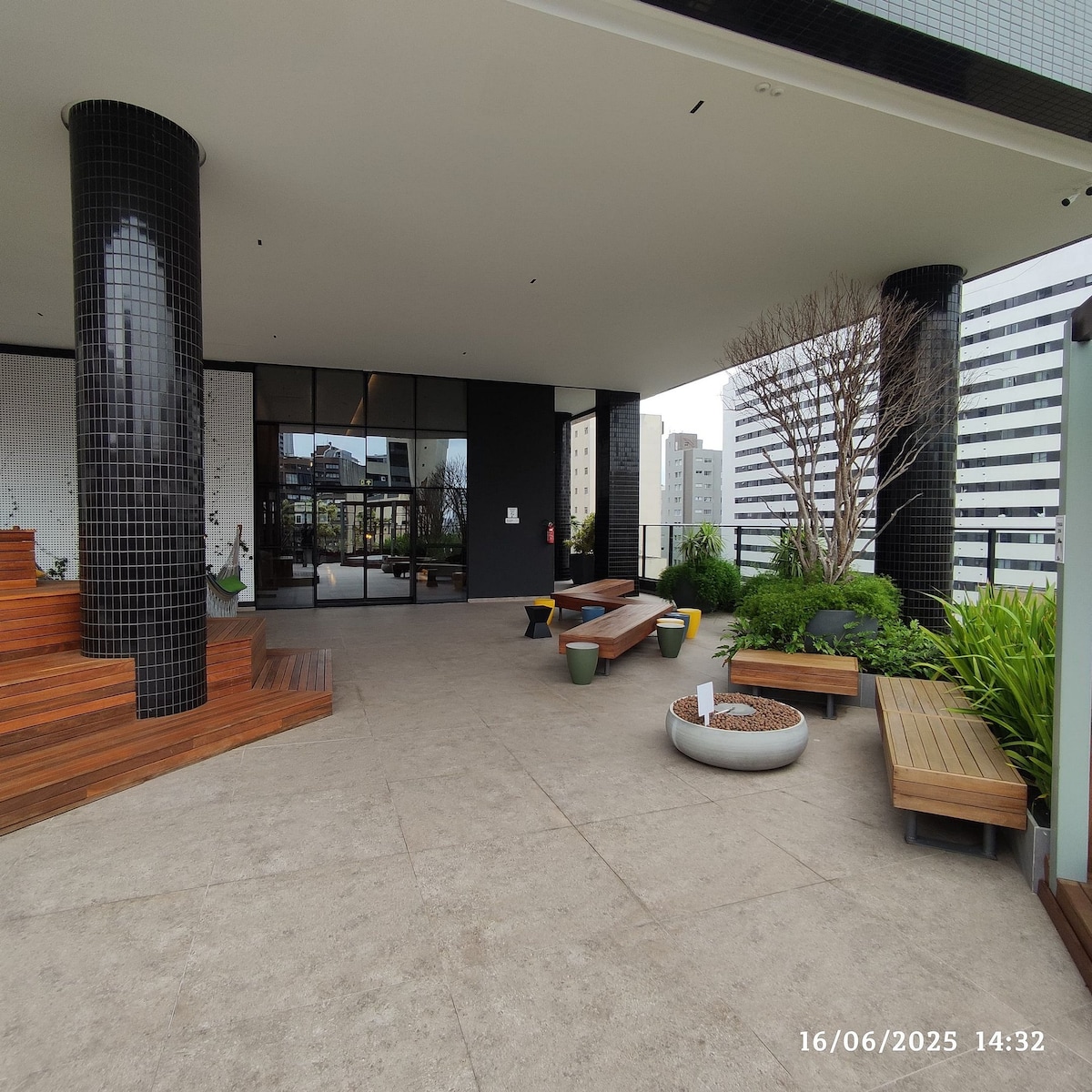 A modern outdoor terrace is featured, surrounded by greenery. Wooden benches offer seating, while potted plants add a fresh touch. Stairs lead to an entrance with large glass doors, allowing views of the city skyline in the background.