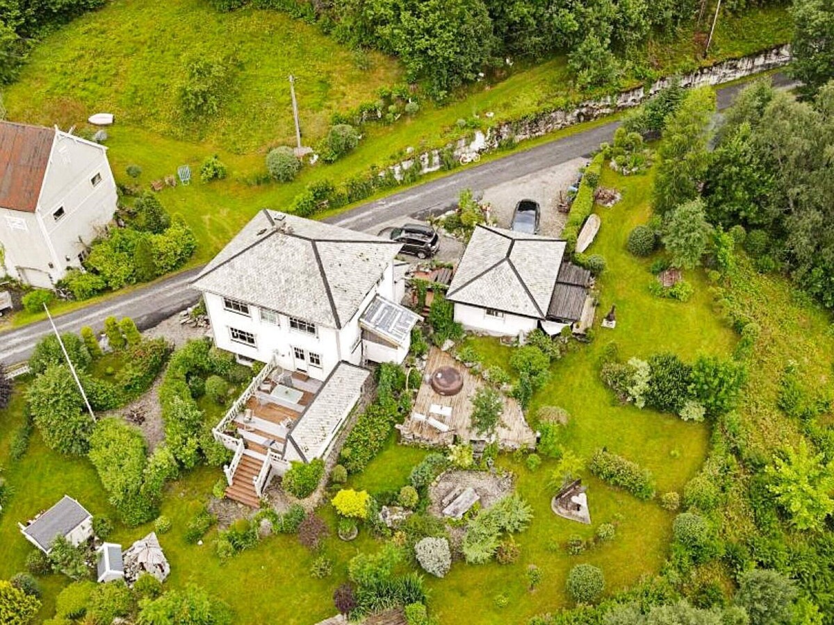 An aerial view of a spacious holiday home set amidst a lush garden. The property includes various green plants and trees, with a distinct terraced area and pathways leading to the house. The surrounding landscape features rolling hills and a small road adjacent to the property.