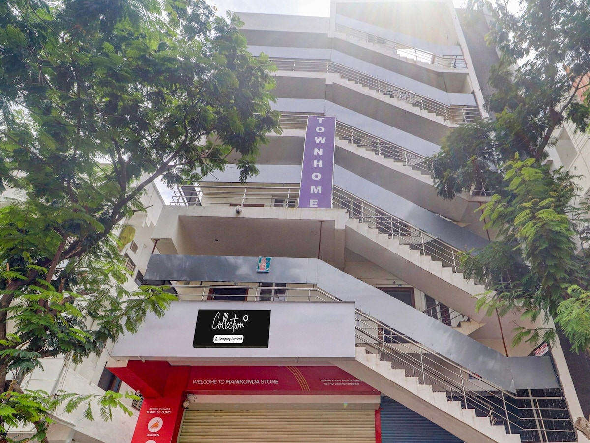 The exterior of a multi-story building is displayed, featuring a modern façade with a staircase leading to the upper floors. Tall trees frame the structure, and a welcoming sign is positioned at the entrance. Bright natural light illuminates the scene.