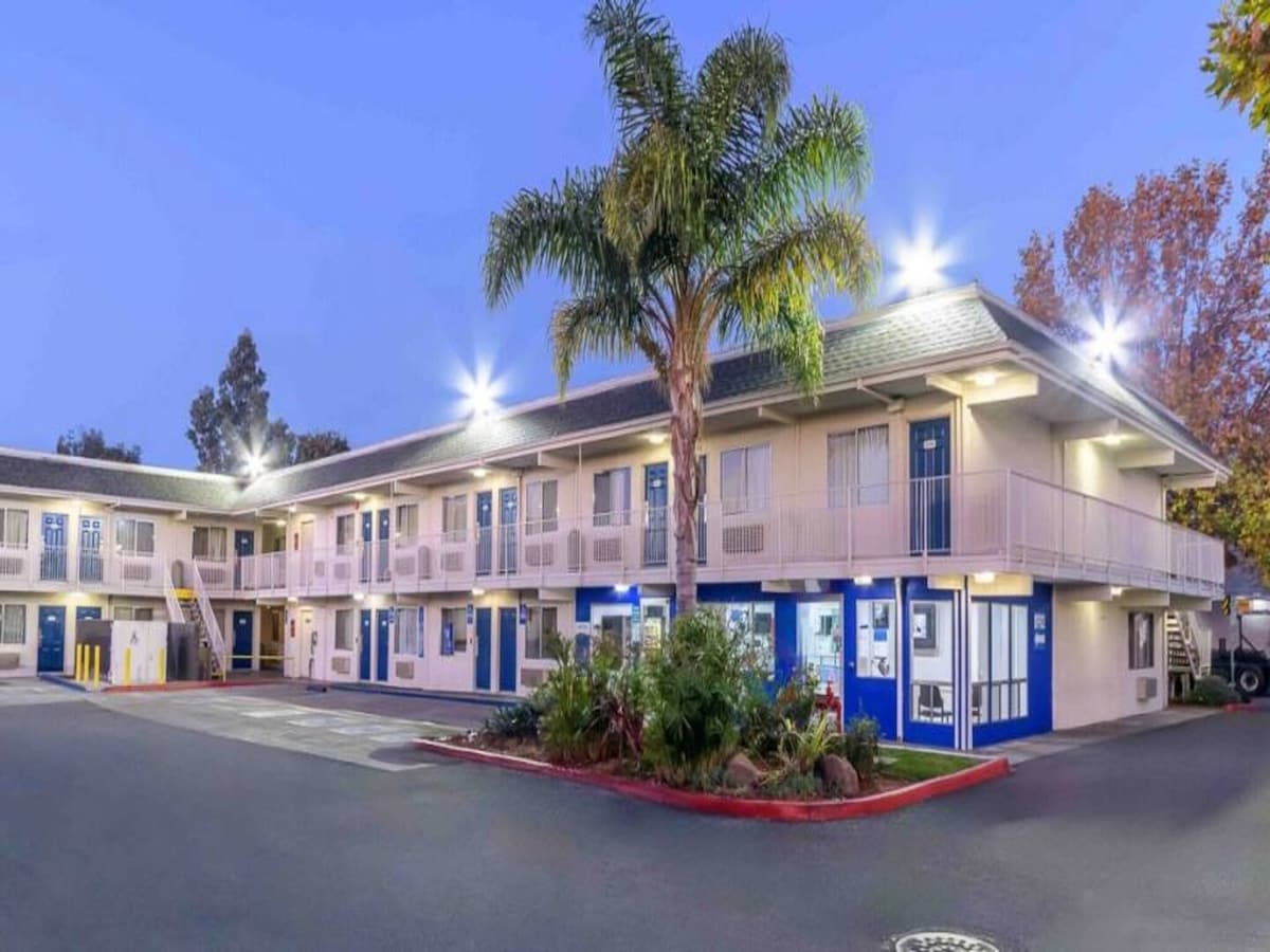 The exterior of the motel is shown, featuring a two-story structure with a balcony lined with railings. Lush greenery and palm trees are visible in the foreground, while bright lights illuminate the building against the twilight sky.