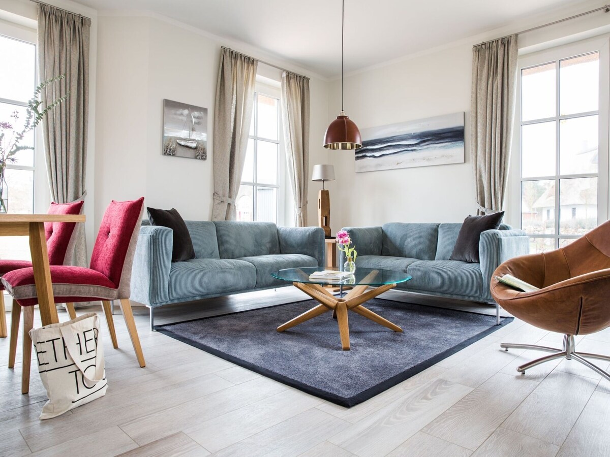 A modern living area features a spacious, light blue sectional sofa paired with a mid-century style coffee table. Natural light filters in through large windows, complemented by elegant curtains. A dining table for four is present, along with a vibrant red chair and a stylish brown armchair.