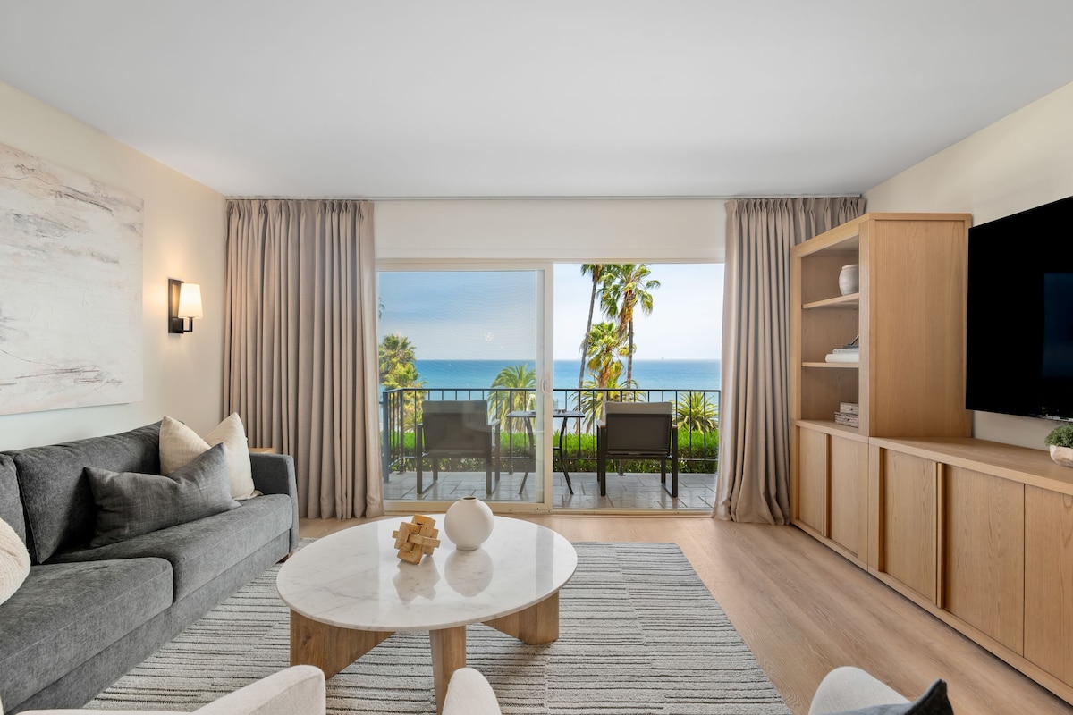 A modern living space features a comfortable gray sofa and a round coffee table atop a textured rug. Floor-to-ceiling glass doors reveal an ocean view and access to a balcony adorned with tropical plants. Soft curtains frame the view, complementing the inviting décor.