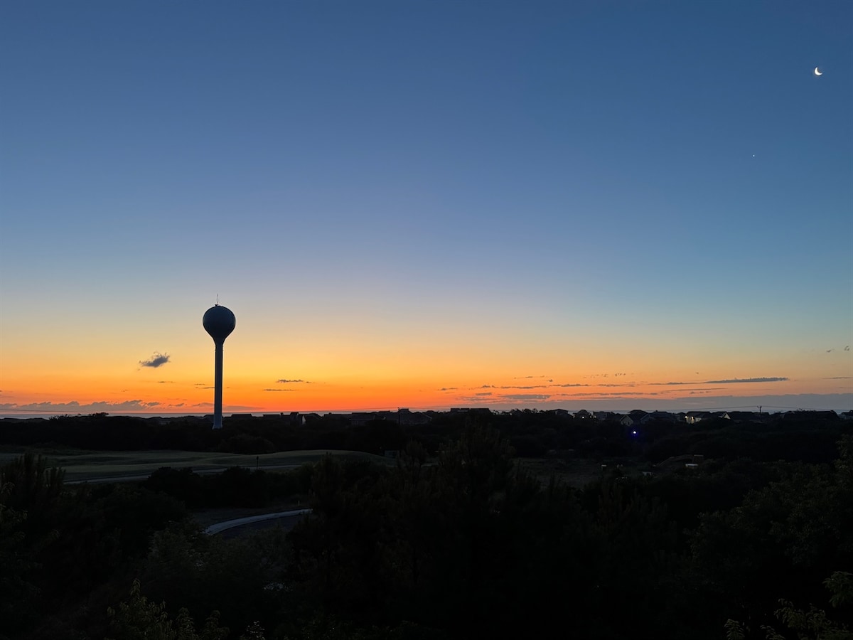 A serene twilight scene captures the vibrant colors of sunset blending into twilight. A tall water tower stands silhouetted against the horizon, while the last light of day reflects off the surrounding landscape, creating a peaceful atmosphere.
