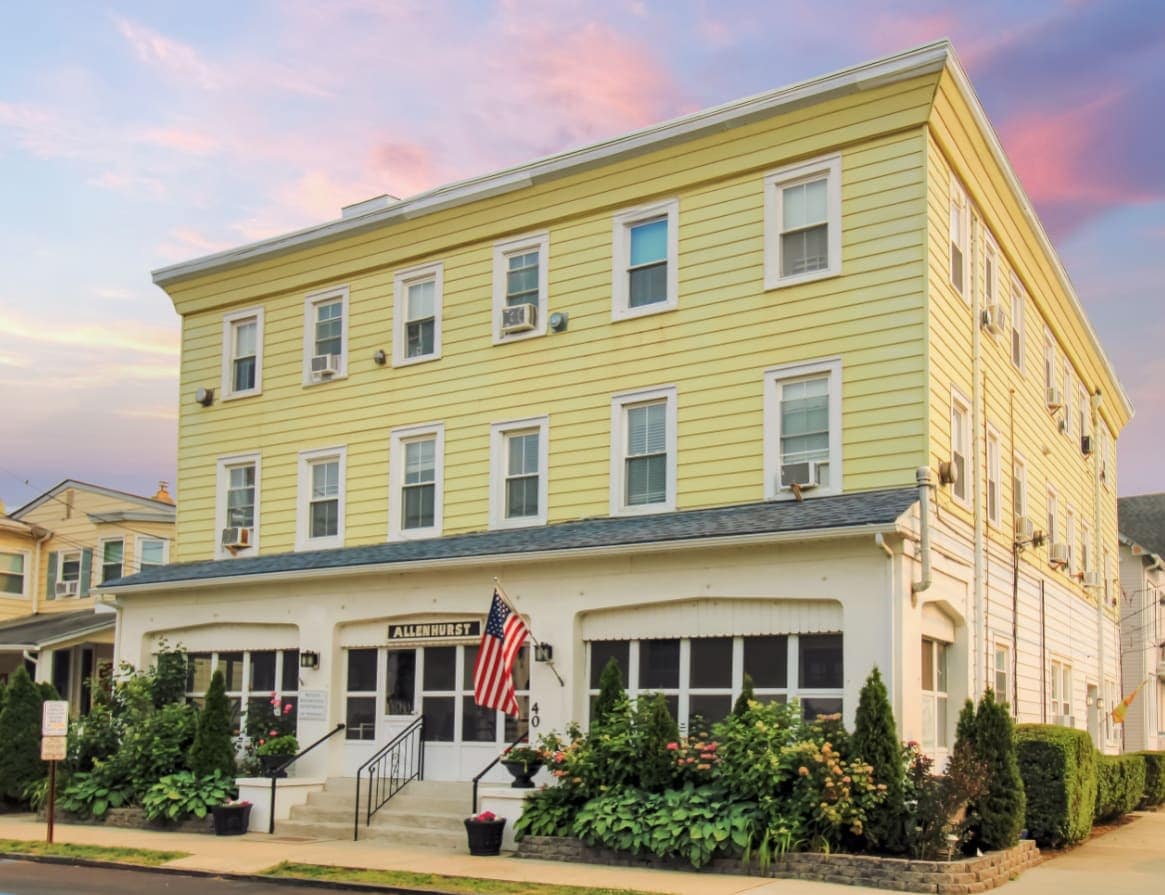 The exterior of The Allenhurst is captured, featuring a three-story building with light yellow siding and white trim. An American flag is displayed in front, surrounded by well-maintained greenery. Large windows provide light to the interior, and a welcoming entrance is accentuated by steps leading to the main door.