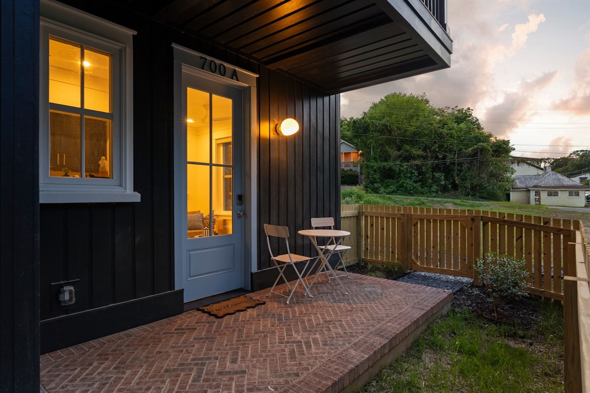 An inviting outdoor space features a brick patio with two foldable chairs and a small table, illuminated by a warm wall light. The entry door, framed by large windows, brightens the entryway, while a fenced yard adds privacy and greenery in the background.
