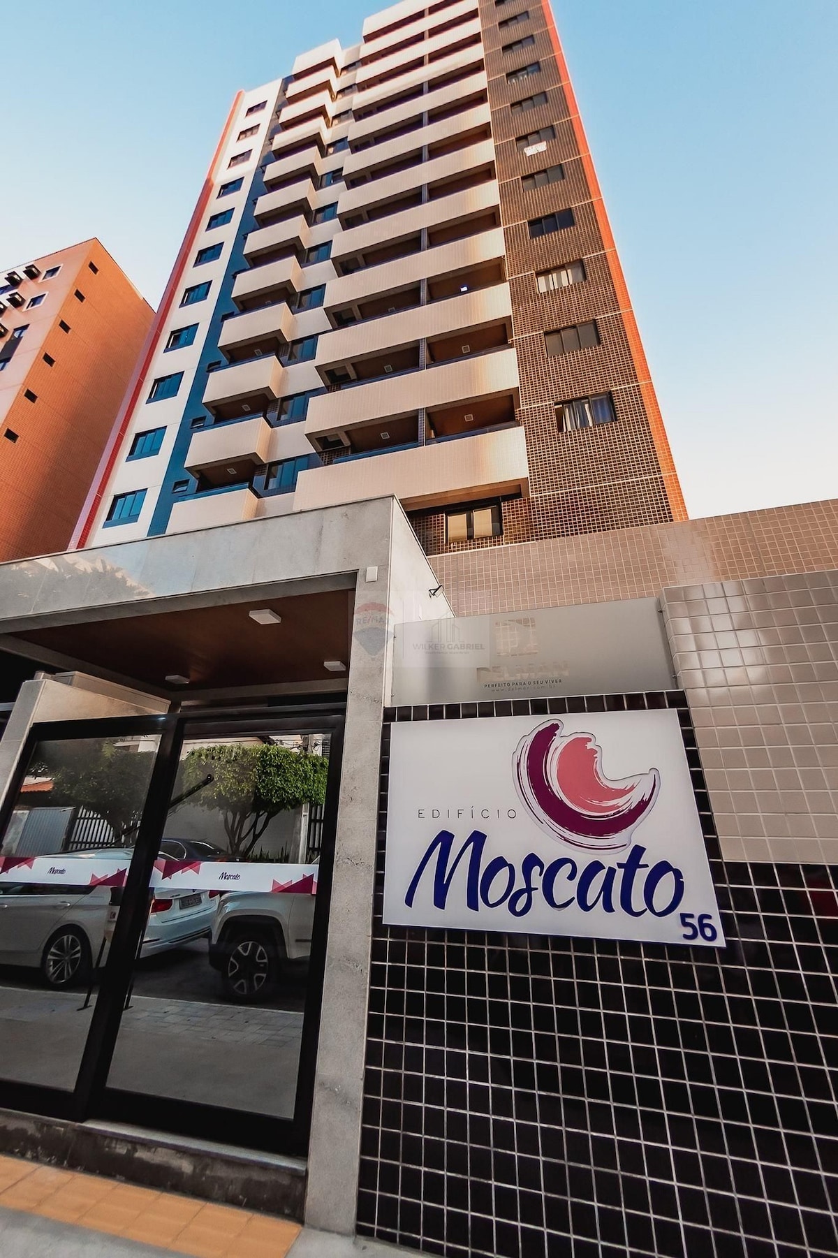 The building facade of Edifício Moscato is displayed, featuring a modern design with distinct blue and red accents. The entrance showcases a clear sign with the building's name. The nearby parking area is visible, with vehicles positioned securely.