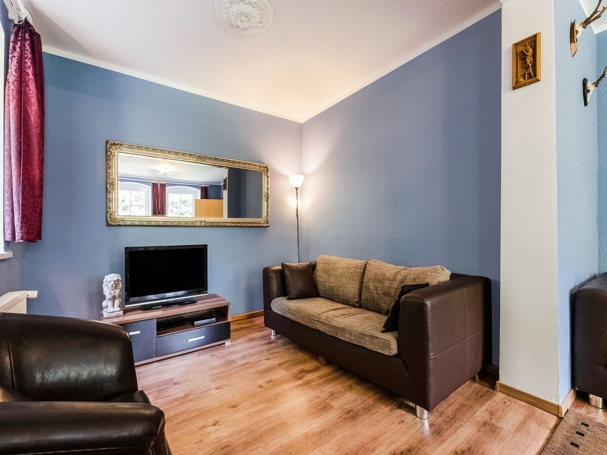 The living room features a comfortable sofa and armchair set against a blue wall. A television is situated on a wooden cabinet, reflecting a simple, modern design. A large mirror enhances the space, while soft lighting creates a relaxed ambiance.