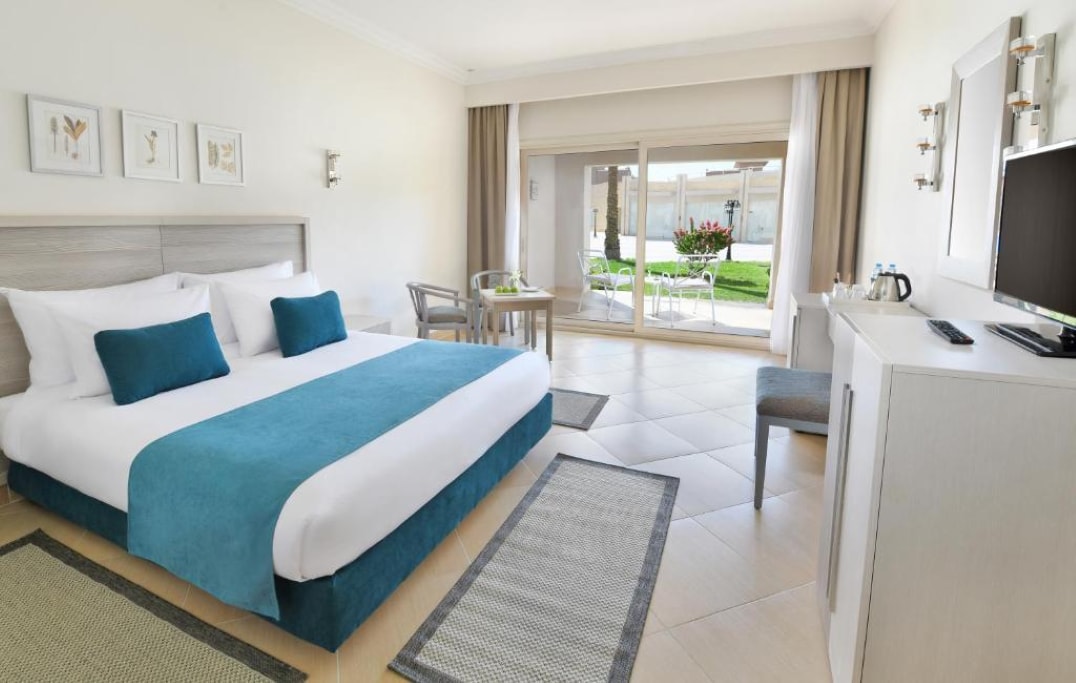 A bright room features a spacious bed dressed in crisp white linens with a teal accent blanket. Natural light filters through large sliding doors opening to a garden. A small dining area includes a table and chairs, and a high definition TV is positioned on a minimalist cabinet.