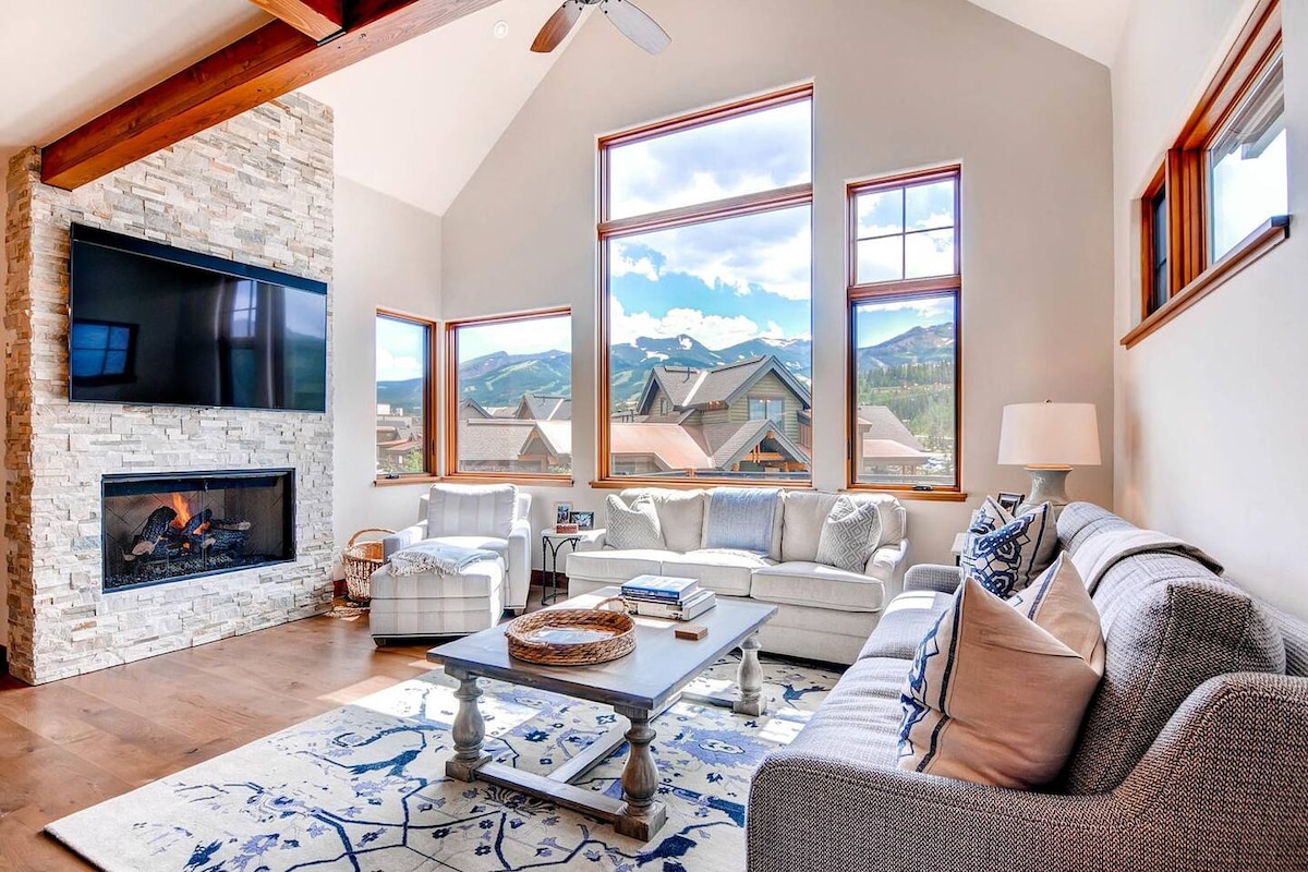 A bright living area is showcased with large windows offering mountain views. A modern stone fireplace serves as a focal point, while a comfortable seating arrangement invites relaxation. The space is enhanced by wooden beams and a patterned area rug that defines the seating area.