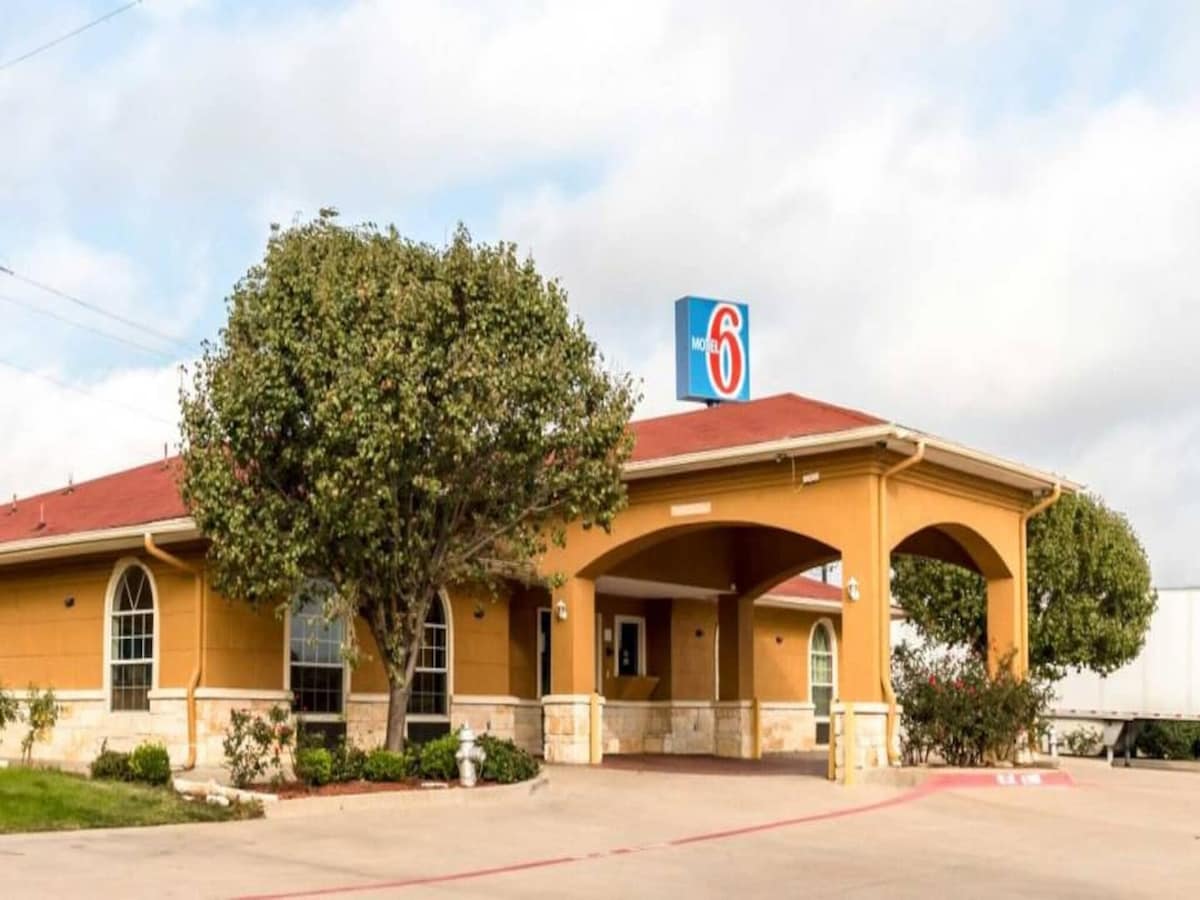 The exterior of the Motel 6 is highlighted by a welcoming entrance with archways and large windows. Landscaping features neatly trimmed bushes and trees, creating an appealing first impression. The prominent motel sign is visible on the roof against a partly cloudy sky.