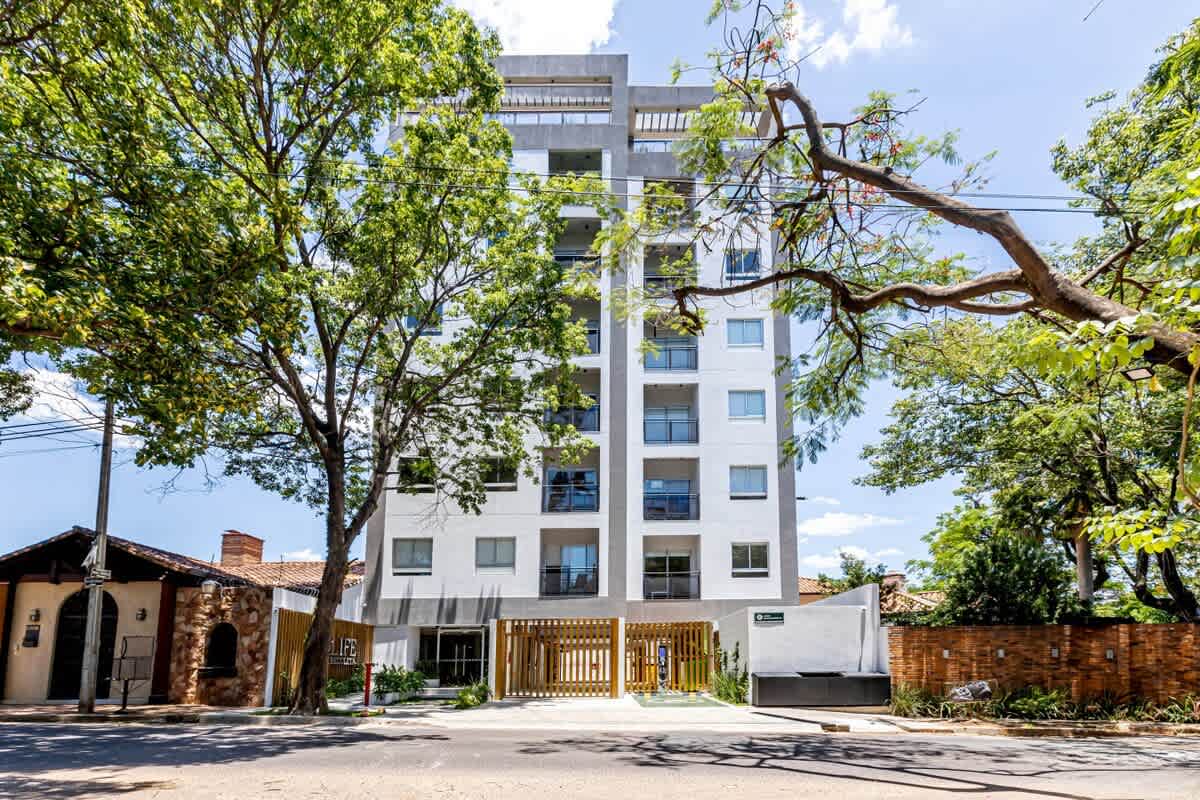 A modern multi-story building is framed by lush trees, creating a serene environment. The structure features a combination of large windows and clean lines, showcasing a contemporary design. The entrance is marked by a decorative gate, adding an element of elegance to the facade.