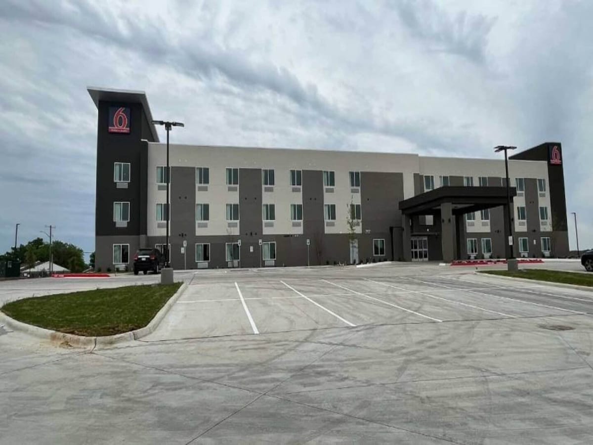The exterior of the hotel is depicted, featuring a modern design with a grey and white color scheme. A covered entrance is visible, along with a spacious parking lot that accommodates multiple vehicles. The sky appears overcast, adding a neutral backdrop.