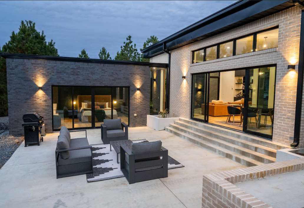 An outdoor patio area is designed with comfortable seating arrangements, featuring a low table and lounge chairs. The modern brick exterior is highlighted by soft outdoor lighting, and large glass doors provide a view into the interior spaces. A grill is also positioned nearby.