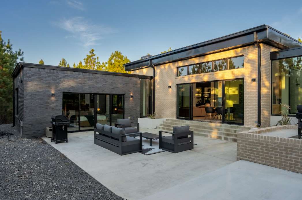 An outdoor patio area features a modern seating arrangement with dark wicker chairs and a low table, complemented by a grill nearby. Large glass doors showcase the interior, allowing glimpses of natural light and views of the surrounding wooded landscape.