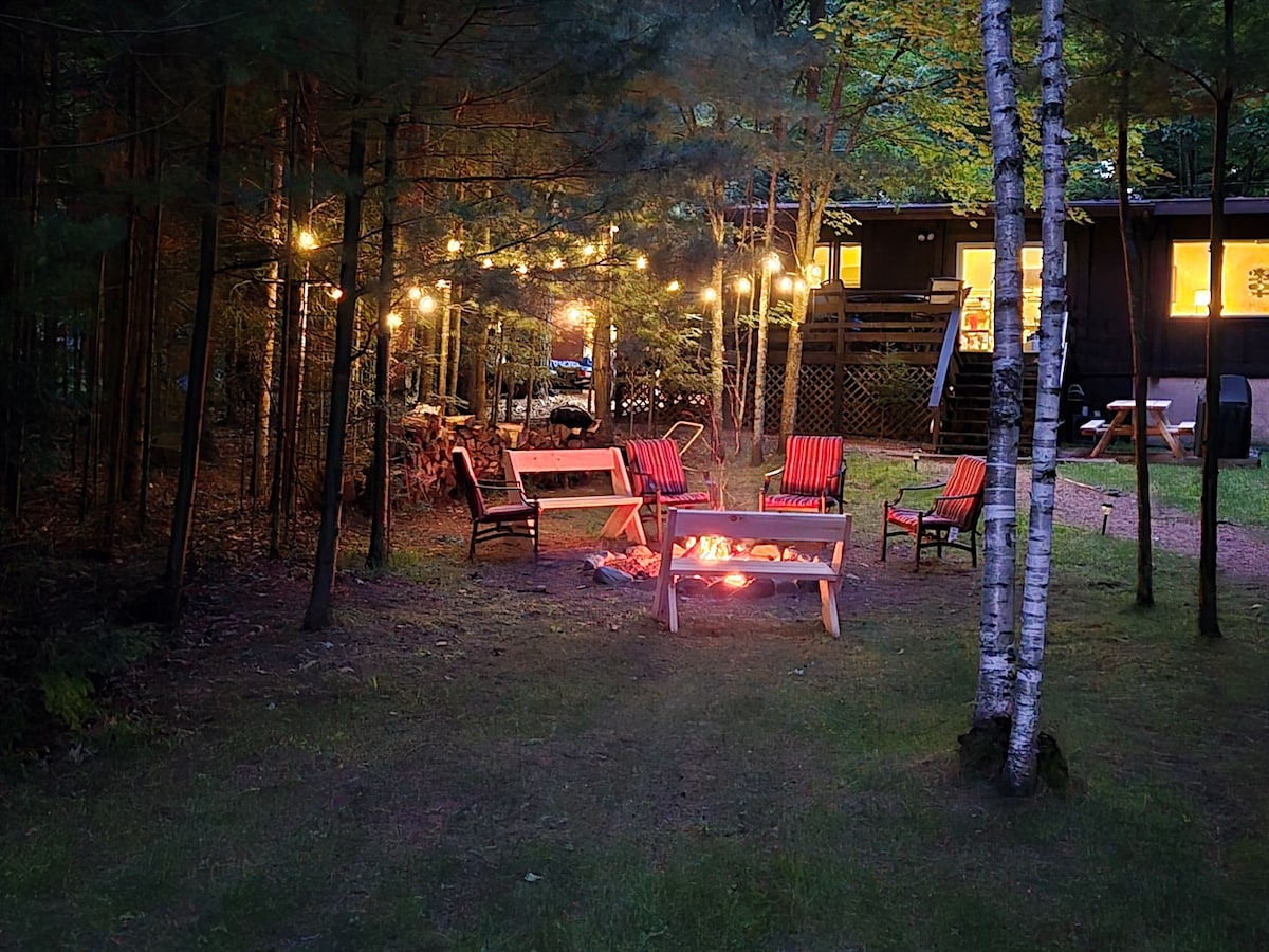 An outdoor fire pit area is surrounded by tall trees, featuring several wooden chairs with bright red cushions. Soft string lights hang above, adding a gentle glow to the scene. A glimpse of the cabin's windows can be seen in the background, inviting warmth from within.