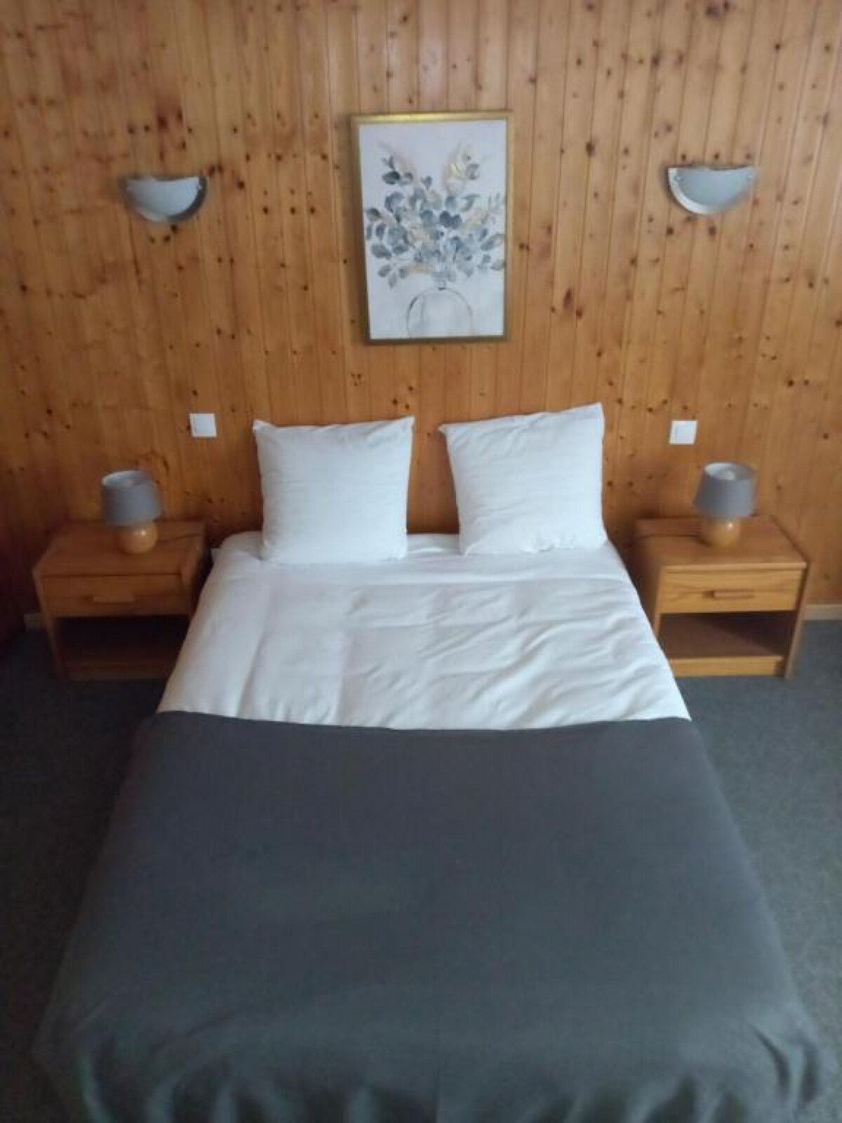 A cozy sleeping area is featured, with a spacious bed dressed in white linens and a gray blanket. Two nightstands with modern lamps are positioned on either side, complemented by a floral wall art piece and warm wooden paneling in the background.