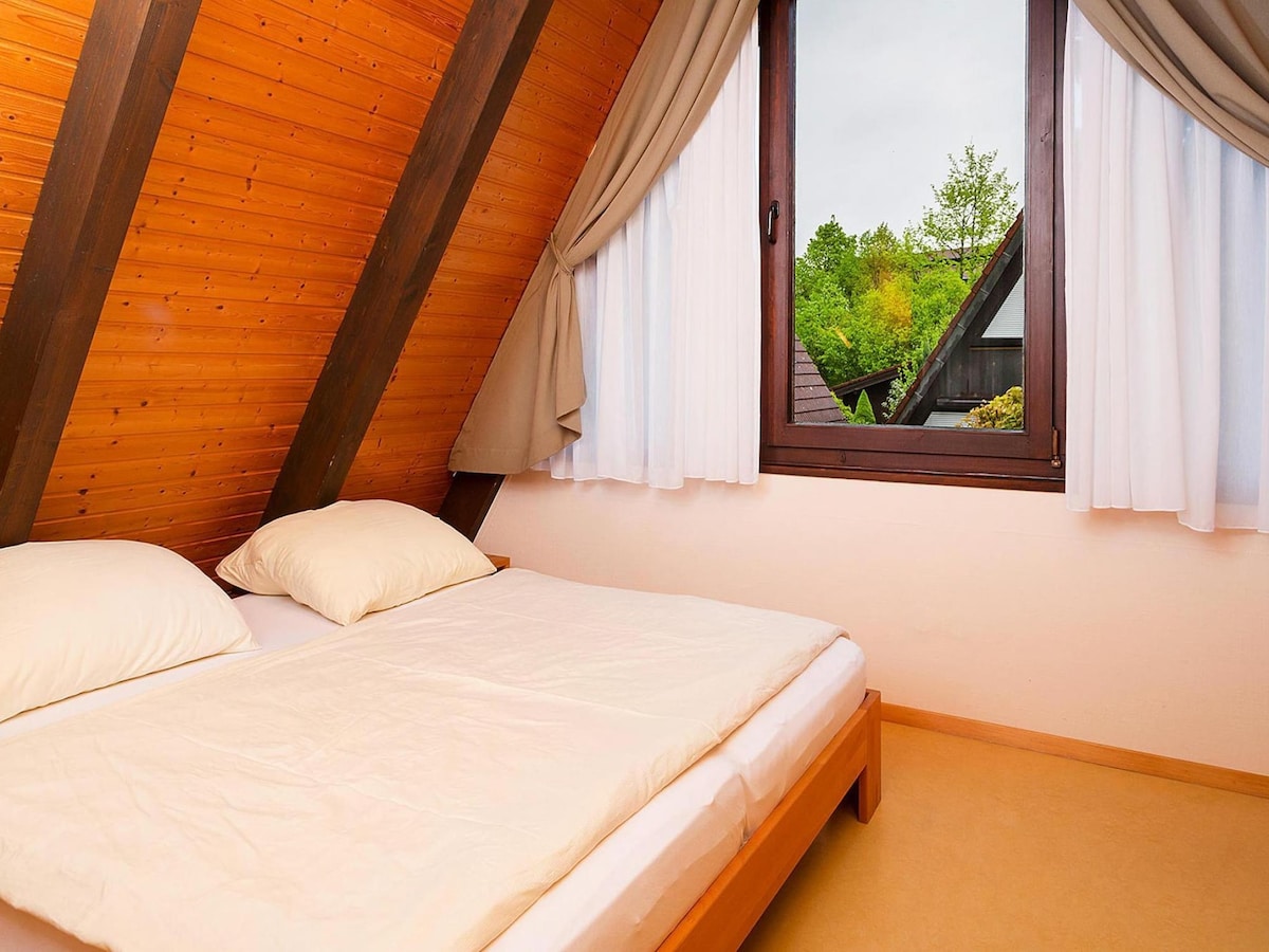 A cozy bedroom features a spacious bed with two soft pillows. Natural light enters through a large window, offering a view of greenery outside. The room is adorned with wooden paneling, creating a warm atmosphere.