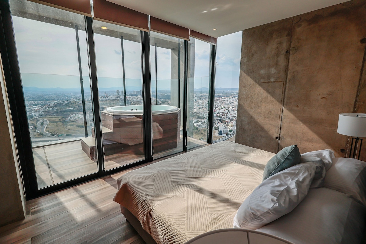A spacious bedroom is featured with a large bed and soft linens. Floor-to-ceiling windows provide spectacular views of the city, with a private jacuzzi positioned nearby. The natural light enhances the modern concrete walls and minimalistic décor, creating a serene atmosphere.