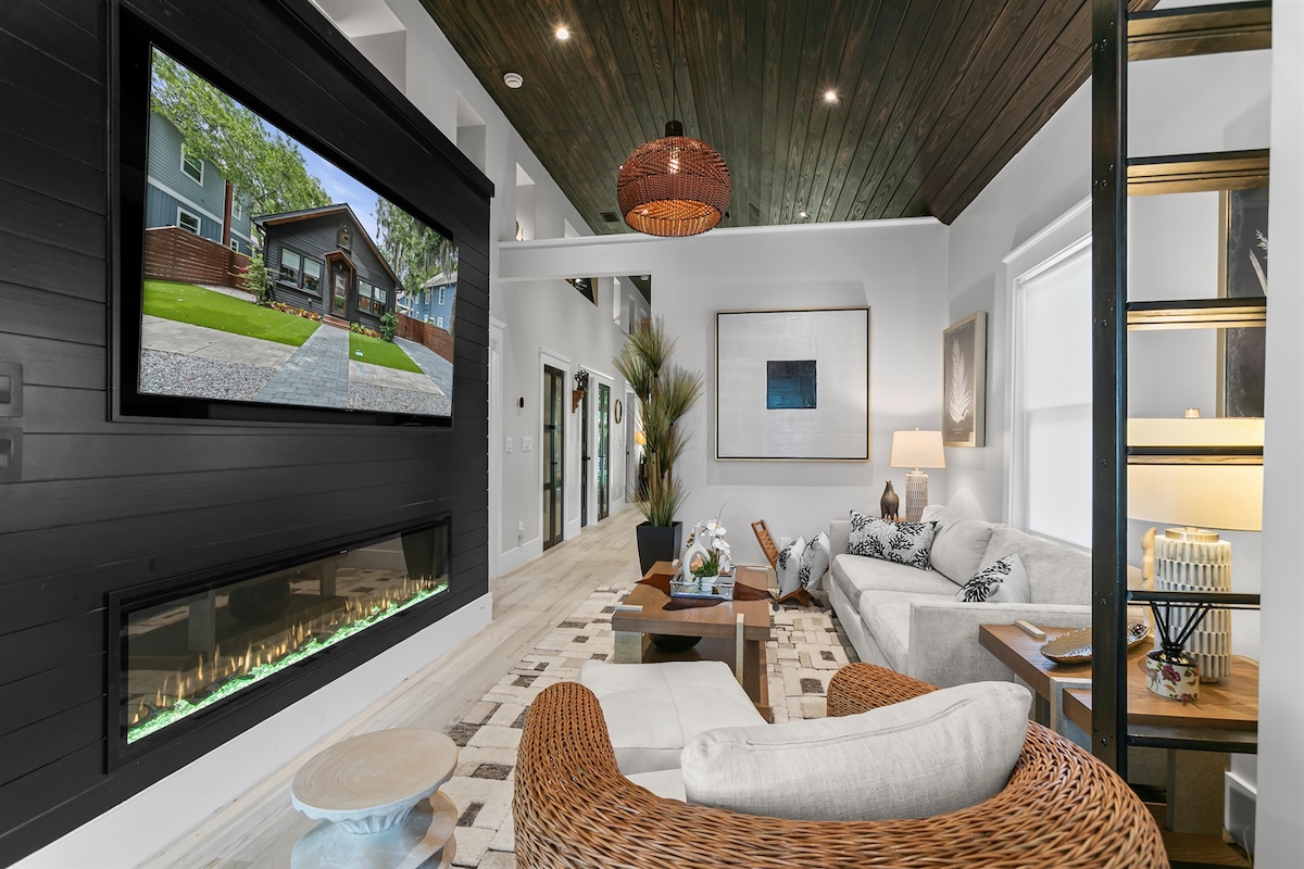 A modern living area highlights a large wall-mounted TV above an electric fireplace. A cozy sectional sofa and a circular chair provide ample seating, while natural light fills the space through large windows. Textured decor and artwork enhance the warm ambiance.