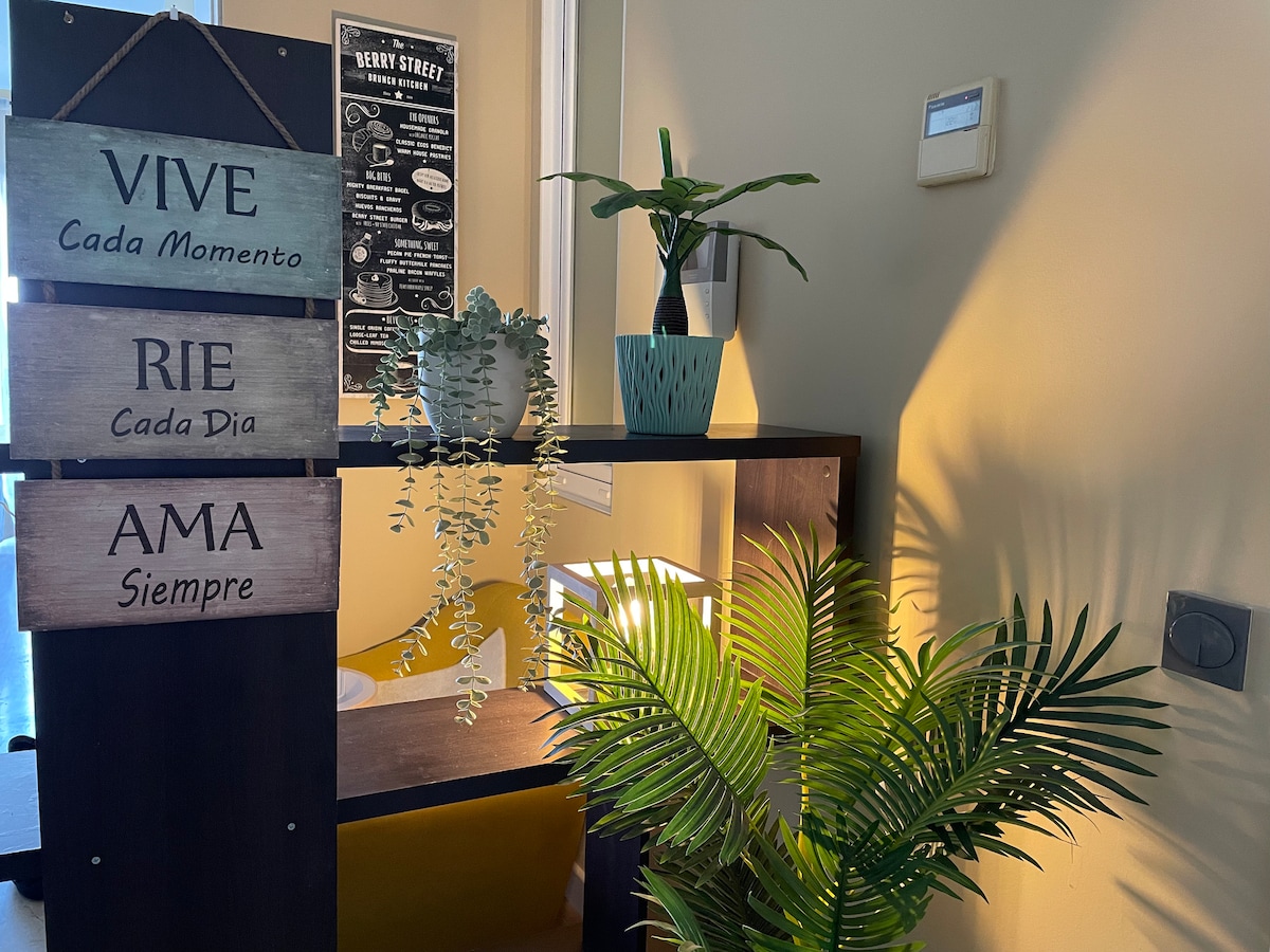 A decorative area is presented with a wooden sign displaying inspiring words in Spanish, surrounded by vibrant green plants. A modern shelf with a potted plant adds a touch of nature, while natural light enhances the inviting atmosphere.