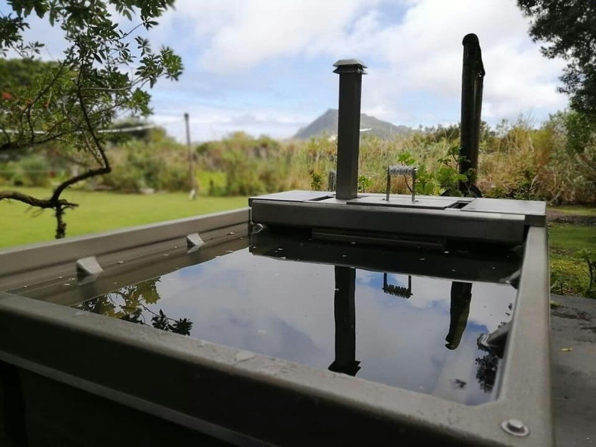 A small braai area with a sleek cooking surface is shown, set against a backdrop of lush greenery and a distant mountain. The reflections on the surface create a serene atmosphere, inviting guests to enjoy outdoor cooking and relaxation.