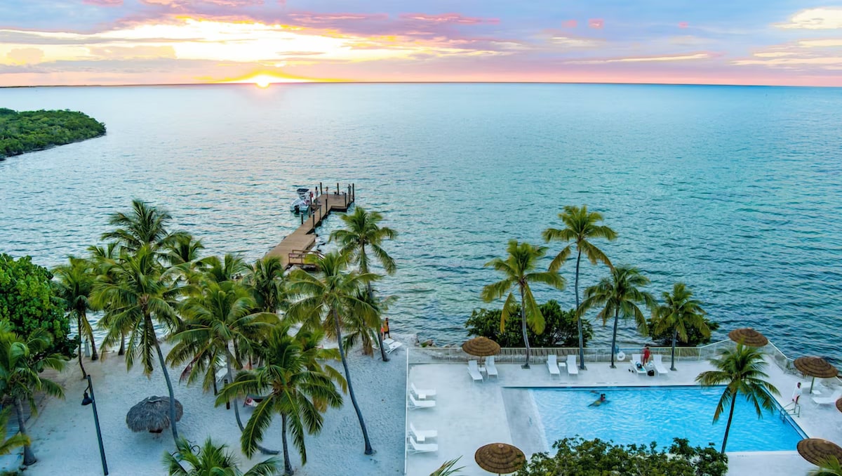A serene waterfront view shows gentle waves lapping against the shore, with palm trees lining the beach area. A clear swimming pool sits in the foreground, with sun loungers positioned around it. A soft sunset blends hues of orange and pink in the sky above.