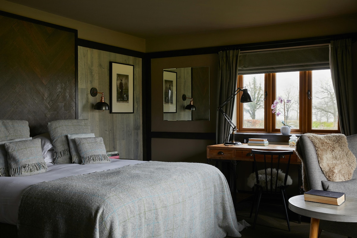 A cozy bedroom is showcased with a spacious bed dressed in neutral linens and a soft throw. A wooden desk with a lamp is nestled by the window, which offers views of the outdoors. The rustic walls are complemented by art and decorative accents.