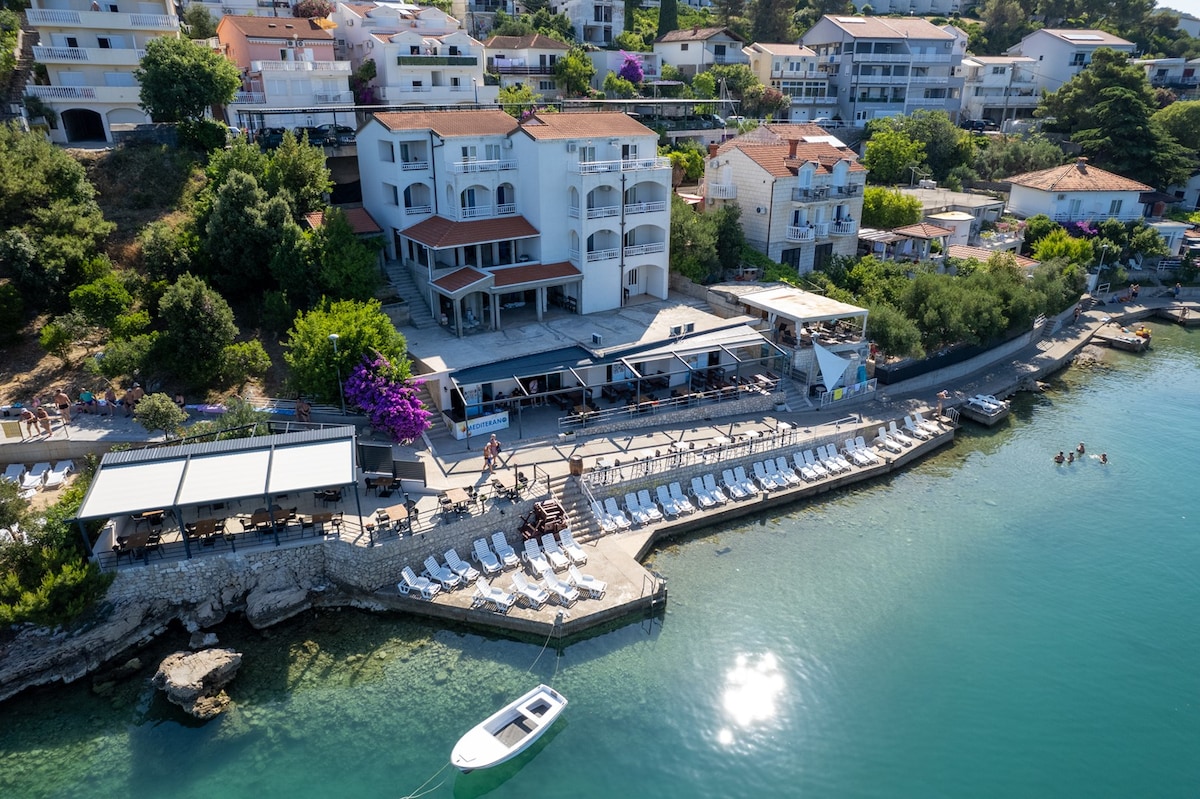 An aerial view captures the property nestled near a calm turquoise bay. The beachfront area features sun loungers arranged on a terrace, with a small boat moored nearby. Lush greenery and blooming flowers enhance the surroundings, complemented by surrounding buildings overlooking the sea.