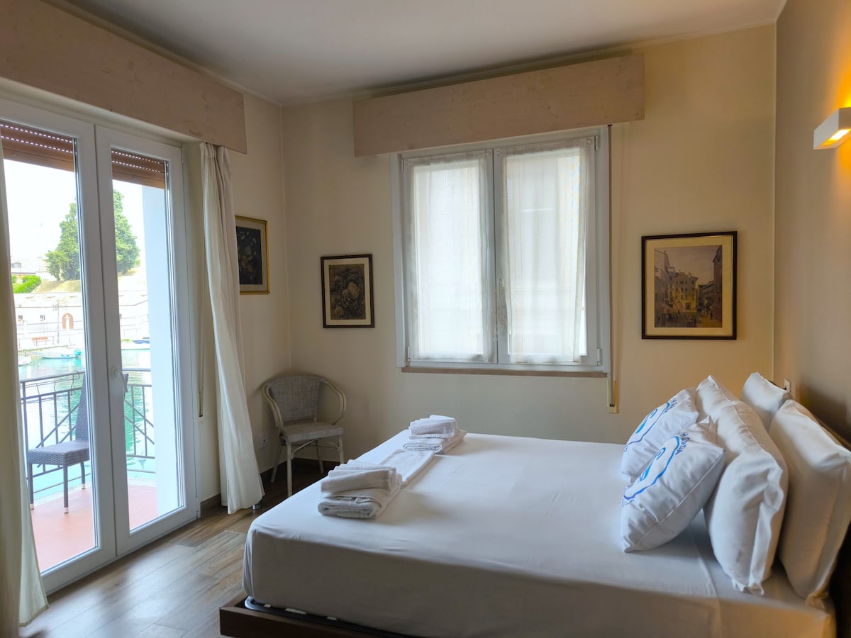 Светла спалня с двойно легло и директен достъп до балкона с прекрасна гледка към канала в Пескера дел Гарда. Изчистените и елегантни мебели създават приветлива и релаксираща атмосфера, идеална за романтичен престой в центъра на града.