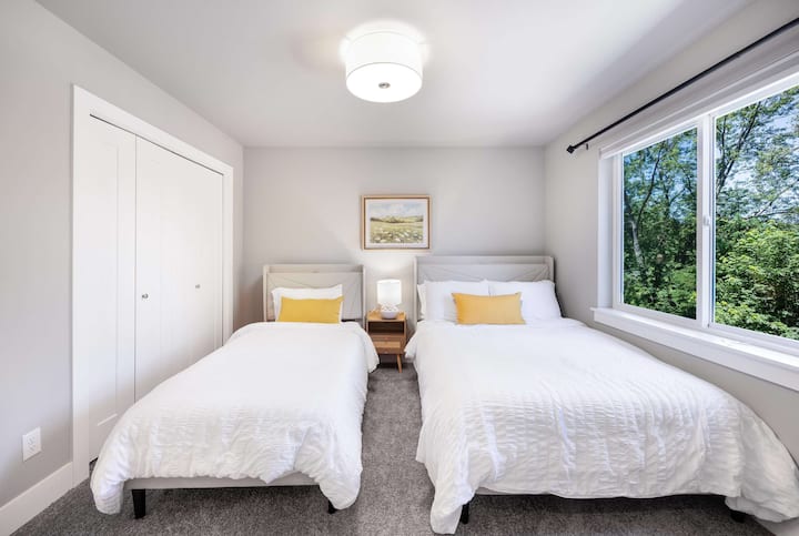 מיטות זוגיות עם כריות צהובות זהות יוצרות סימטריה במרחב המשותף הזה. שידת הלילה מעץ בין המיטות מציעה מקום אחסון משותף.