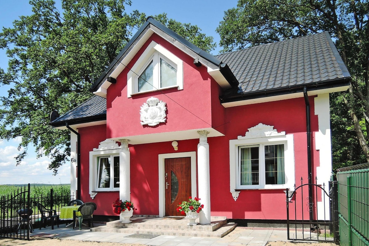The holiday home showcases a vibrant red exterior with white accents and decorative columns. Large windows provide ample natural light, and flower pots are positioned at the entrance. A well-maintained garden area complements the charming structure, set against a backdrop of greenery.