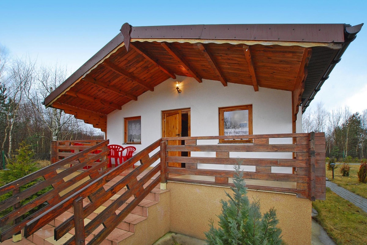 A cozy holiday home is displayed with a wooden sloped roof and a covered terrace featuring two red chairs. A staircase leads to the entrance, while a railing provides safety. The surrounding area includes greenery and sparse trees, lending a sense of serenity.