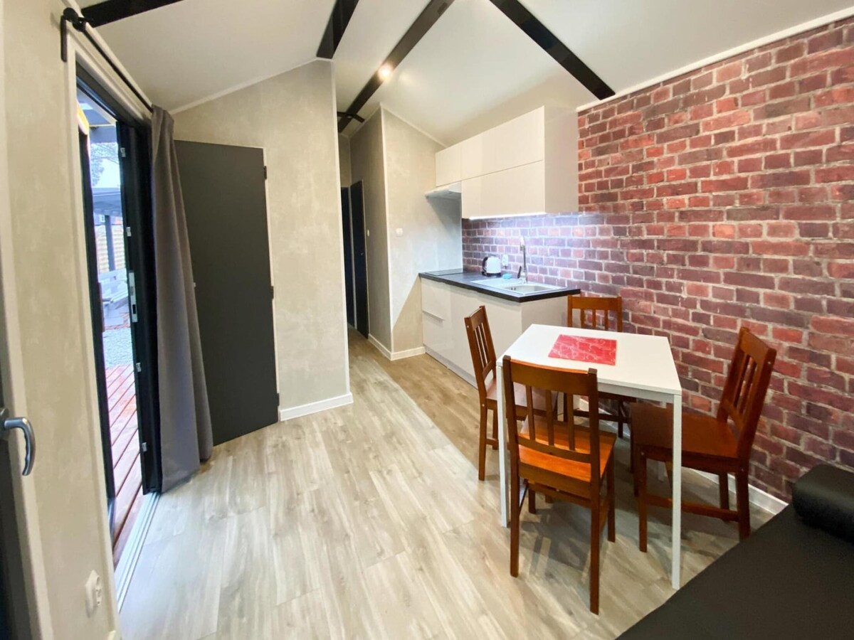 A modern interior showcases an open-plan living space with a dining table and chairs. Exposed brick walls contrast with sleek cabinetry in the kitchenette, creating a cozy atmosphere. Natural light filters in through the sliding door, which leads to an outdoor area.