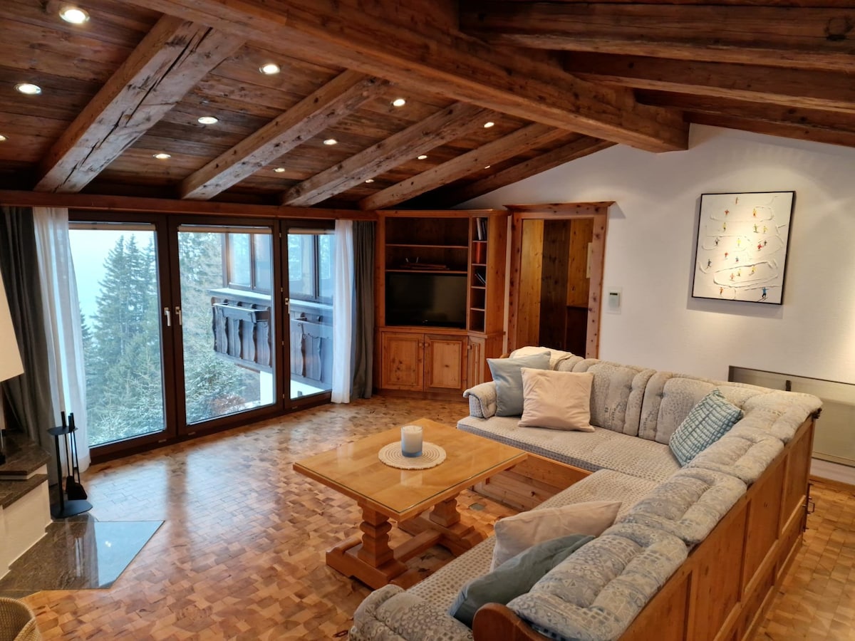 The living area features a spacious wooden ceiling with exposed beams, enhancing the alpine charm. A sectional sofa surrounds a central wooden table, providing seating for relaxation. Large windows offer views of the surrounding trees, allowing natural light to fill the room.