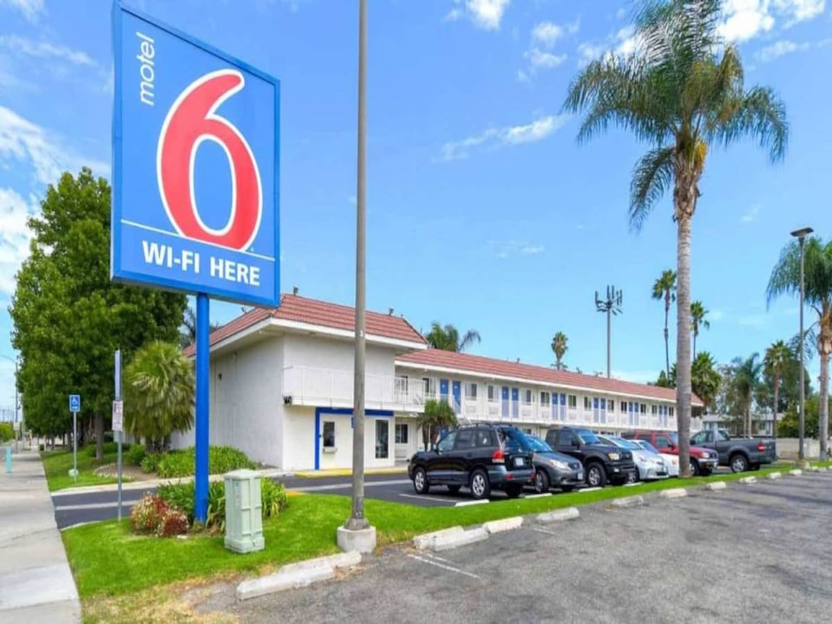 The exterior of a motel is shown, featuring a prominent sign displaying 'Motel 6' and 'Wi-Fi Here.' A parking area is visible with several parked cars and palm trees lining the premises, set against a clear blue sky.