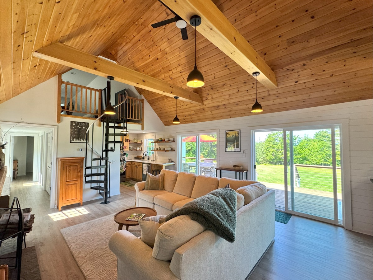 A cozy living space features a light-filled area with a large, corner couch and natural wood paneling on the ceiling. A spiral staircase leads to a loft above. Large sliding doors open to an inviting deck overlooking green outdoor spaces.